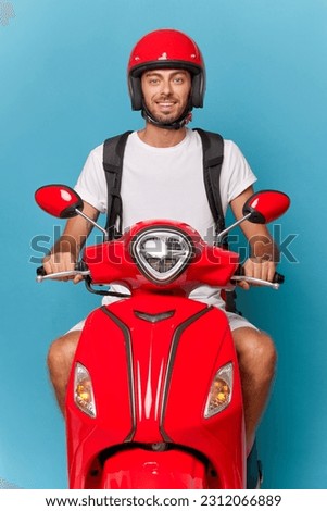 Young tourist guy riding red scooter, wearing white t-shirt and red helmet, holds both hand on handle bar looking ahead, nice trip concept, copy space