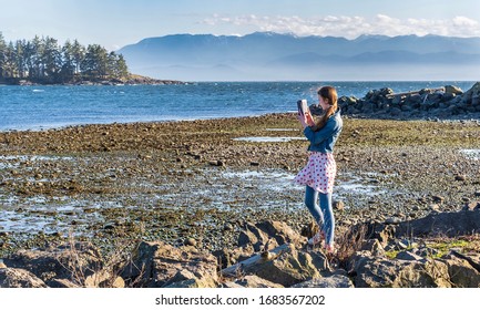 Young Tourist Takes Cell Phone Photo Of Rugged Ocean Scenery With Hazy Mountains On A Bright Sunny Spring Day  - In Sooke, Vancouver Island, British Columbia, Canada