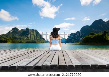 Young tourist wearing a blue shirt and black hat. Sitting on an old wooden bridge looking at the beautiful view of the lake, mountains, in summer. She is happy to travel and relax on weekends.