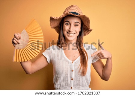 Young tourist woman on vacation wearing summer hat holding hand fan over yellow background with surprise face pointing finger to himself