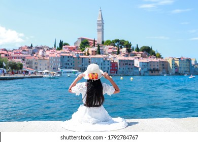Young tourist woman sitting on Harbour holding her hat and looking to beautiful cityscape of rovinj city, Croatia in the summer. Happy freedom girl traveling in Europe she wearing white dress.