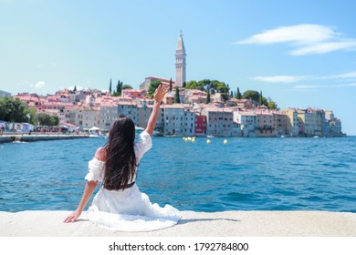 Young tourist woman sitting on Harbour holding her hand up wave to beautiful rovinj city, Croatia. Happy traveler girl enjoy on vacation in Europe. Freedom lady with white dress on a sunny day.