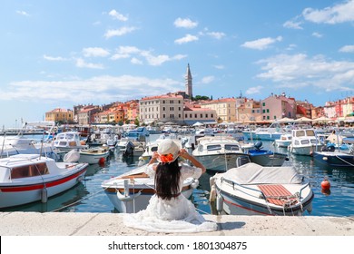 Young tourist woman sitting on harbor holding her hat and looking to beautiful rovinj city in the summer.Happy freedom Asian girl traveling in Croatia, Europe. Lady on vacation at the sea and blue sky