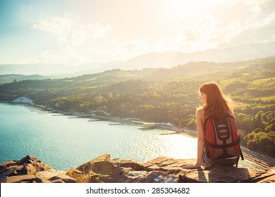 Young tourist woman is sitting on the top of the mounting and looking at a beautiful sea bay landscape. Hiking woman with backpack relaxing on the top of the cliff enjoying sunlit sea view.  