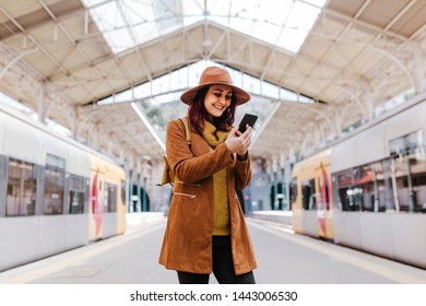 Young Tourist Woman At Train Station Waiting To Take A Train And Travel. Using Mobile Phone And Smiling. Tourism And Lifestyle Concept