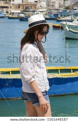 Young tourist woman in white straw hat enjoying a view of the bay with lot small colorful fishing boats, a summertime vacation concept in a charming seaside town