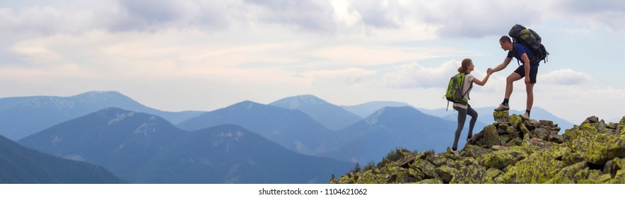 Young tourists with backpacks, athletic boy helps slim girl to clime rocky mountain top against bright summer sky and mountain range background. Tourism, traveling and healthy lifestyle concept. - Shutterstock ID 1104621062