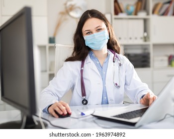 Young Trainee Doctor In A Protective Mask, Undergoing An Internship At A Clinic During A Pandemic, Sits At A Desktop ..in Front Of A Computer In Office