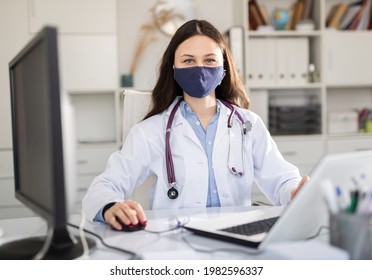 Young Trainee Doctor In A Protective Mask, Undergoing An Internship At A Clinic During A Pandemic, Sits At A Desktop ..in Front Of A Computer In Office