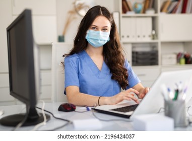 Young Trainee Doctor In A Protective Mask, Undergoing An Internship At A Clinic During A Pandemic, Sits At A Desktop ..in Front Of A Computer In Office