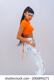 A Young Trap Dancer With Braids. Black Breed Girl Of African Ethnicity With Orange T-shirt And Cowboy Pants On A Gray Background, Treft Posé