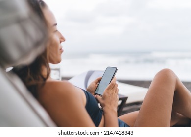Young travel blogger resting in recreation lounge zone using digital mobile technology for web networking, millennial woman in swimwear lying at sunbed connecting to 4g wireless on cellular device