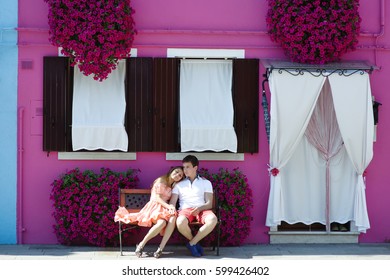 Young Travel Couple On Vacation Relaxing On A Bench Near A Purple House In Burano, Venice, Italy