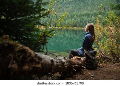 Young Travel Woman Siting On Deadwood Near Amazing Lake On Coniferous Forest Background. Concept Of Mountains Trip. Side View. Meditation In Nature. Relax. Travel Journal. Fantastic Weekends.