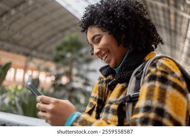 Young traveler with a backpack smiling while using a smartphone in a spacious, modern train station. The scene conveys connection, exploration, and communication in a vibrant setting - Powered by Shutterstock