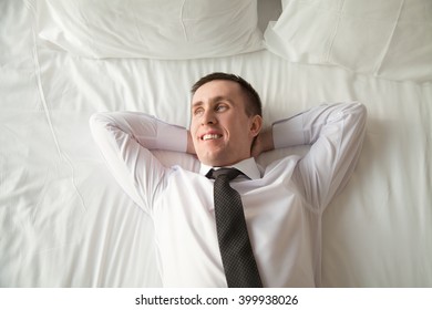 Young Traveler Businessman Wearing White Shirt And Necktie Lying On The Bed In The Hotel Room With His Hands Behind Head. Office Worker Relaxing After Coming Home With Blissful Expression