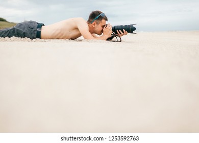 Young Traveler Man Lying On Beach Near Sea On Vacation And Making Photos Of Nature. Topless Handsome Barefoot Man With Camera And Big Lense Spying People From Afar. Time For Hobby. Boy Resting On Sand