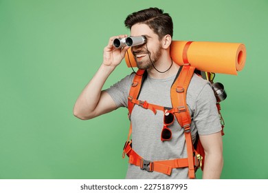 Young traveler white man carry backpack stuff mat watch in binocular isolated on plain green background. Tourist leads active healthy lifestyle walk on spare time. Hiking trek rest travel trip concept