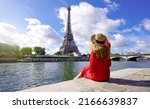 Young traveler woman in red dress and hat sitting on the quay of Seine River looking at Eiffel Tower, famous landmark and travel destination in Paris.