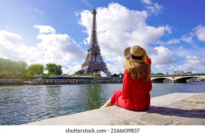 Young traveler woman in red dress and hat sitting on the quay of Seine River looking at Eiffel Tower, famous landmark and travel destination in Paris.