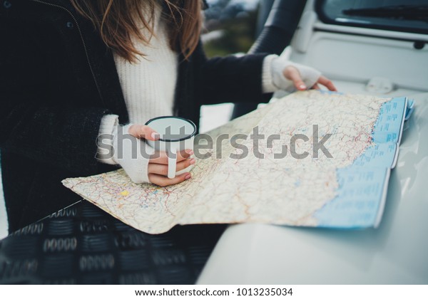 Young traveler woman standing outdoor with\
touristic map exploring countryside, lifestyle travel concept,\
hipster girl enjoying active vacation in mountains looking on map\
and drinking tea outdoor