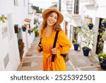 A young traveler woman in a vibrant orange dress and hat enjoys a sunny stroll through a charming white-washed village with flower pots. Concept of lifestyle, vacation, fashion, travel