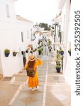 A young traveler woman in a vibrant orange dress and hat enjoys a sunny stroll through a charming white-washed village with flower pots. Concept of lifestyle, vacation, fashion, travel