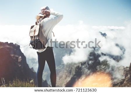 Young traveler woman wearing backpack and hat standing on edge of cliff and looking at clouds, sunset and mountains.