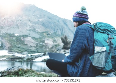 Young traveling girl with backpack and hat sitting on rock and using laptop to connect network in the stunning mountain wilderness in front of amazing cold lake.