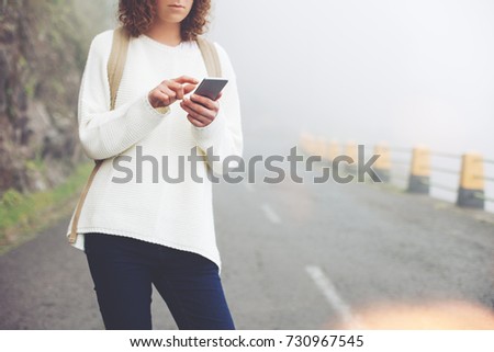 Young traveling girl with backpack and hat standing on the road in fog, searching path and using mobile phone.