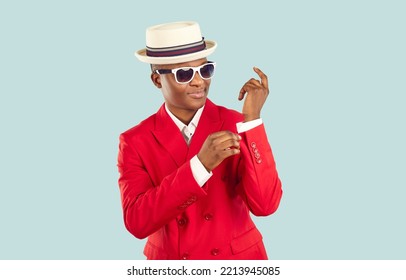 Young trendy African American man in red suit and hat for attending party or fashionable popular event, adjusts sleeves and prepares for work of showman or presenter standing in pastel blue studio - Shutterstock ID 2213945085