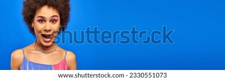 Young and trendy african american woman with bold makeup and natural hair sticking out tongue and looking at camera isolated on blue, charismatic model in summer outfit, banner