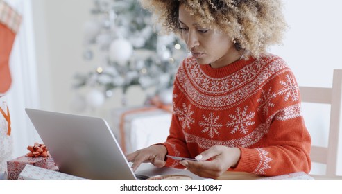 Young trendy African woman doing Xmas shopping online sitting in front of the decorated Christmas tree at home entering her card details on the computer