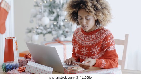 Young trendy African woman doing Xmas shopping online sitting in front of the decorated Christmas tree at home entering her card details on the computer