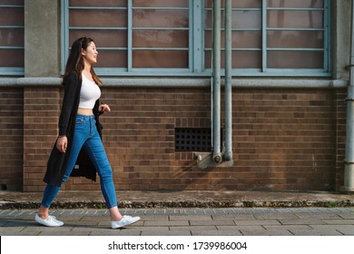 Young Trendy Asian Chinese Woman Wearing Blue Skinny Jeans And Crop Top Shirts Walking On Street In Summer Time. Side View Full Length Girl With Sunglasses On Head Walking By Red Brick Wall Enjoy Sun