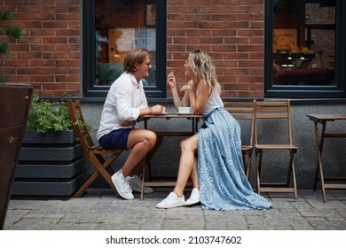 Young Trendy Couple Talking And Smile At Coffee Shop On Street Of City. Stylish Man And Woman In Love Full Length Portrait At Restaurant. Date At Cafe Table Outdoors Coffeehouse