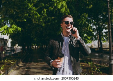 Young Trendy Fun Smiling Man In Black Leather Jacket Eyeglasses Walk In Green Park Hold Takeaway Craft Paper Cup Drink Coffee To Go Talk By Mobile Cell Phone Concept Of Urban Lifestyle Spring Season