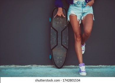 Young trendy girl with a surf skateboard in pink sneakers and stylish blue jeans shorts is standing in front of grey wall background and showing shaka sign. Active and healthy lifestyle concept.