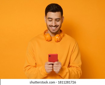 Young Trendy Man Wearing Sweatshirt And Wireless Headphones, Smiling While Looking At Content On Phone Screen, Isolated On Orange Background