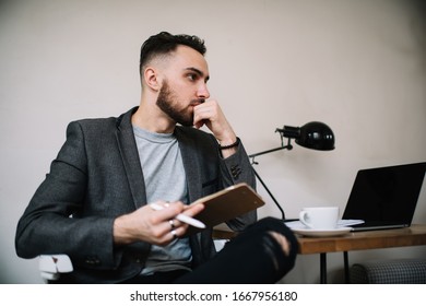 Young Trendy Thoughtful Writer Inspiring By Ideas Over Cup Of Coffee At Work Desk With Laptop Sitting In Ripped Jeans And Holding Clothespin Tablet  Touching Beard And Looking Away