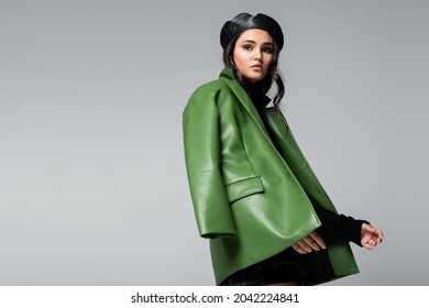 young trendy woman in black beret and green leather jacket posing isolated on grey
