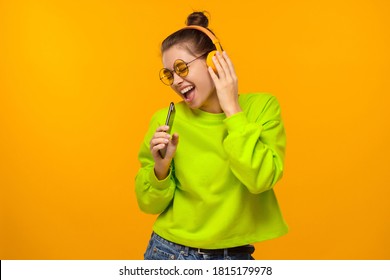 Young Trendy Woman Singing With Closed Eyes, Holding Phone As Microphone, Isolated On Yellow Background