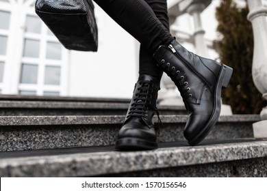 Young trendy woman in stylish leather black boots in jeans with a bag stands on the stone steps in the city. Close up of female legs in fashion footwear. New collection of shoes and accessories.