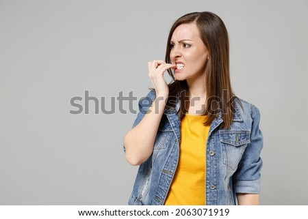 Young troubled pensive puzzled indignant caucasian woman 20s wear casual denim jacket yellow t-shirt look aside biting nails isolated on grey color background studio portrait People lifestyle concept