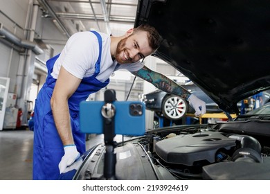 Young Troubleshooter Technician Car Mechanic Man In Blue Overalls White T-shirt Talk Mobile Cell On Phone Stand Show Process Fix Problem With Raised Hood Work In Vehicle Repair Shop Workshop Indoors.