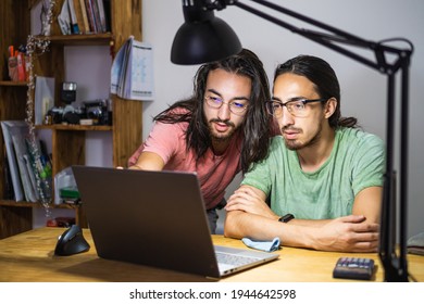 young twin brothers with long hair and glasses looking at computer, FreeLancer work with family at home, gray laptop with vertical wireless mouse, dramatic light with architect's lamp, library