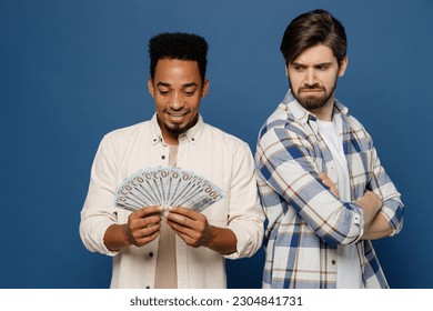 Young two friends envious grumpy men 20s wear white casual shirts together hold in hand fan of cash money in dollar banknotes isolated plain dark royal navy blue background. People lifestyle concept
