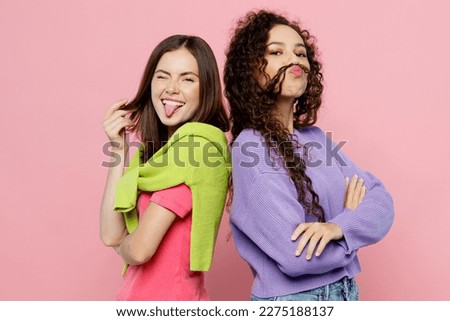 Young two friends funky women wears green purple shirts hold hands crossed folded stand back to back show tongue make fake hair mustache together isolated on pastel plain light pink color background