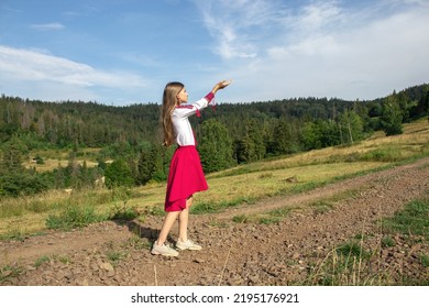 A young Ukrainian girl in a national suit stands in a meadow in the mountains, looking at the sky and asking for heaven, hands raised to the sky. Independence Day.