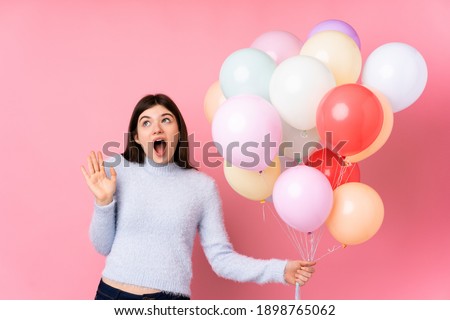 Young Ukrainian teenager girl holding lots of balloons over isolated pink background with surprise facial expression